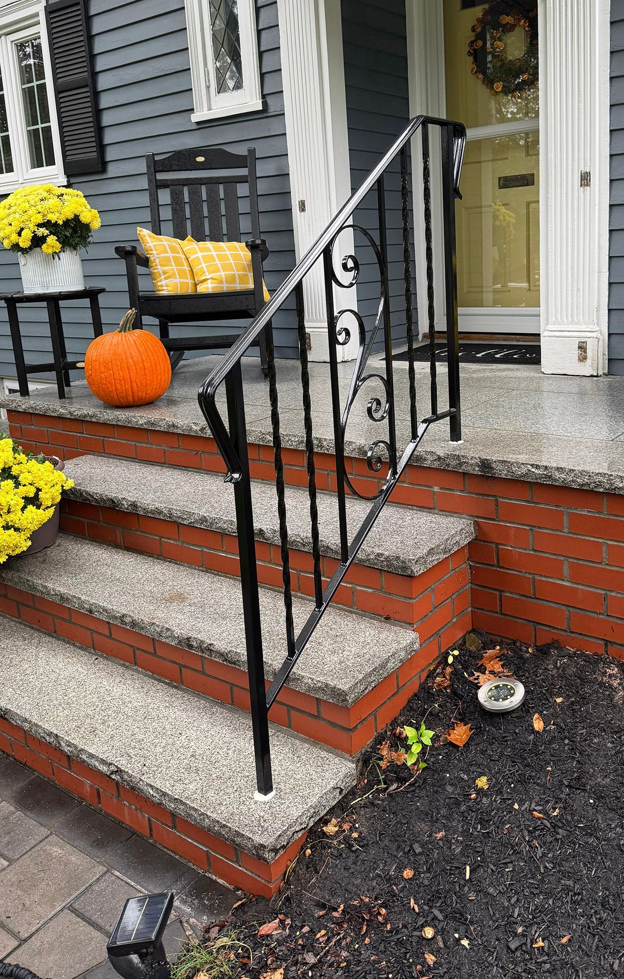 Front porch with autumn decor, brick steps, black railing, and yellow flowers beside a gray house