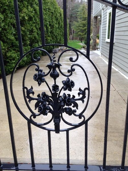 Black metal gate featuring a central circular design with decorative floral scrollwork, set against a driveway and house.