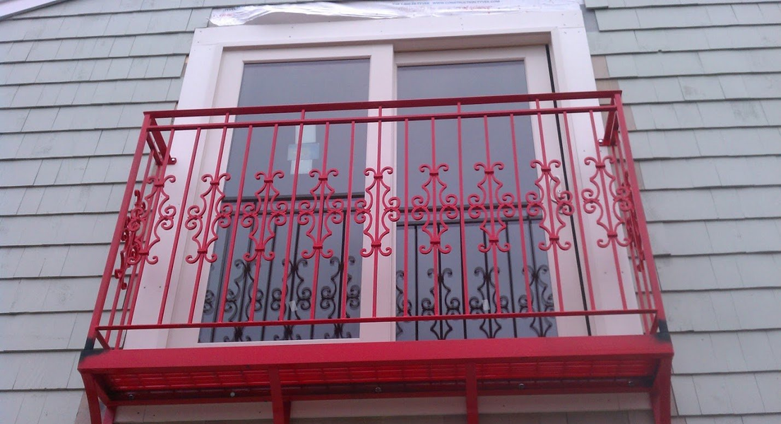 A bright red, decorative wrought-iron balcony attached to the exterior of a building with light blue siding.