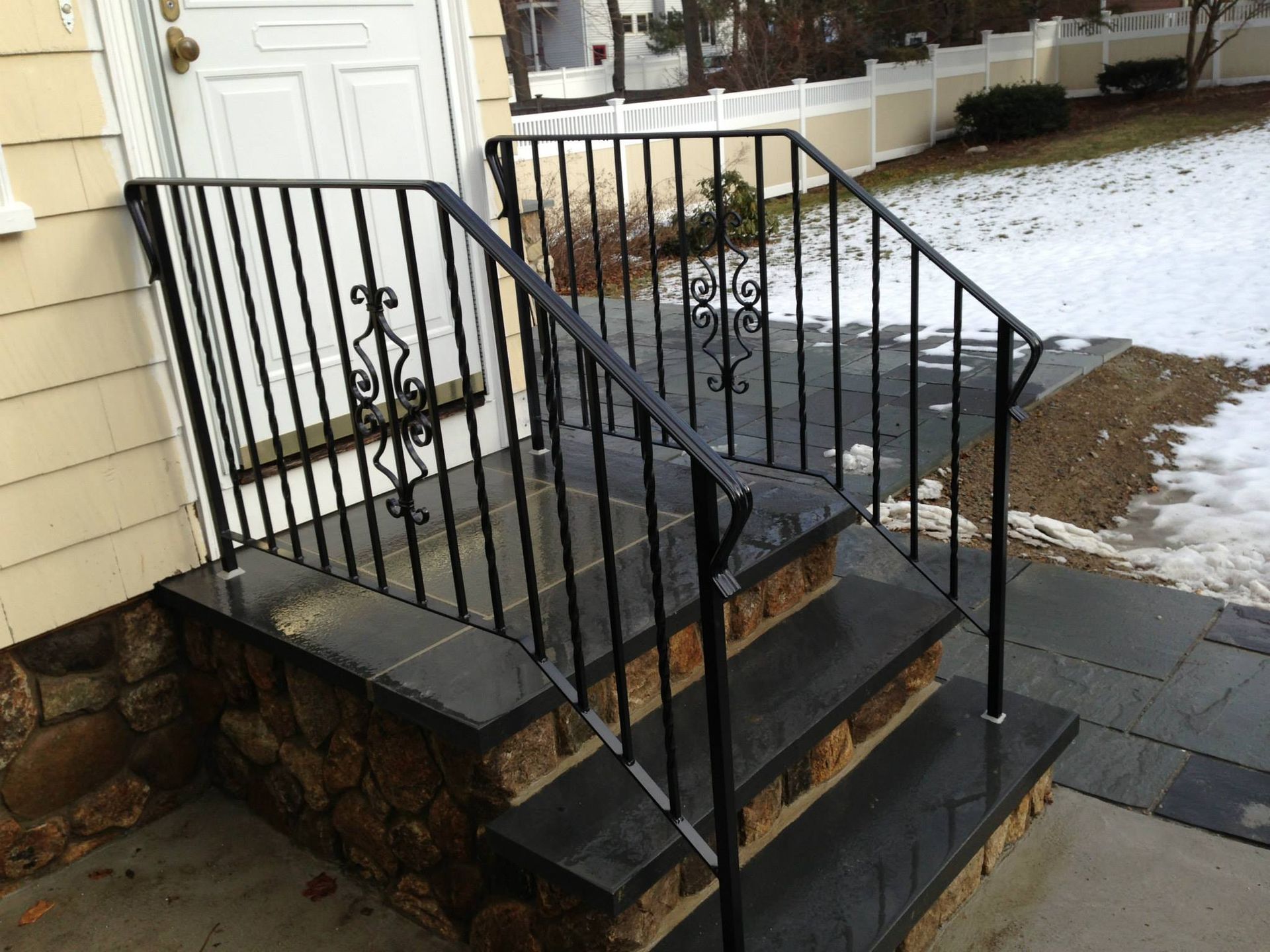 Snow-dusted front steps with black metal railings beside a white door and stone walkway