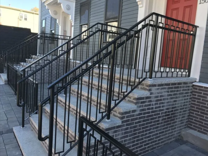 Black metal handrails lining a front staircase beside a brick stoop and red door