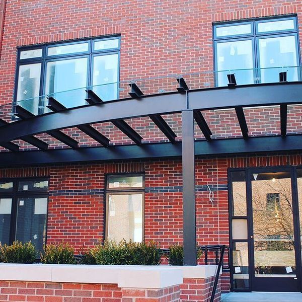 A modern brick building entrance features a large black metal pergola structure over a glass door and windows.