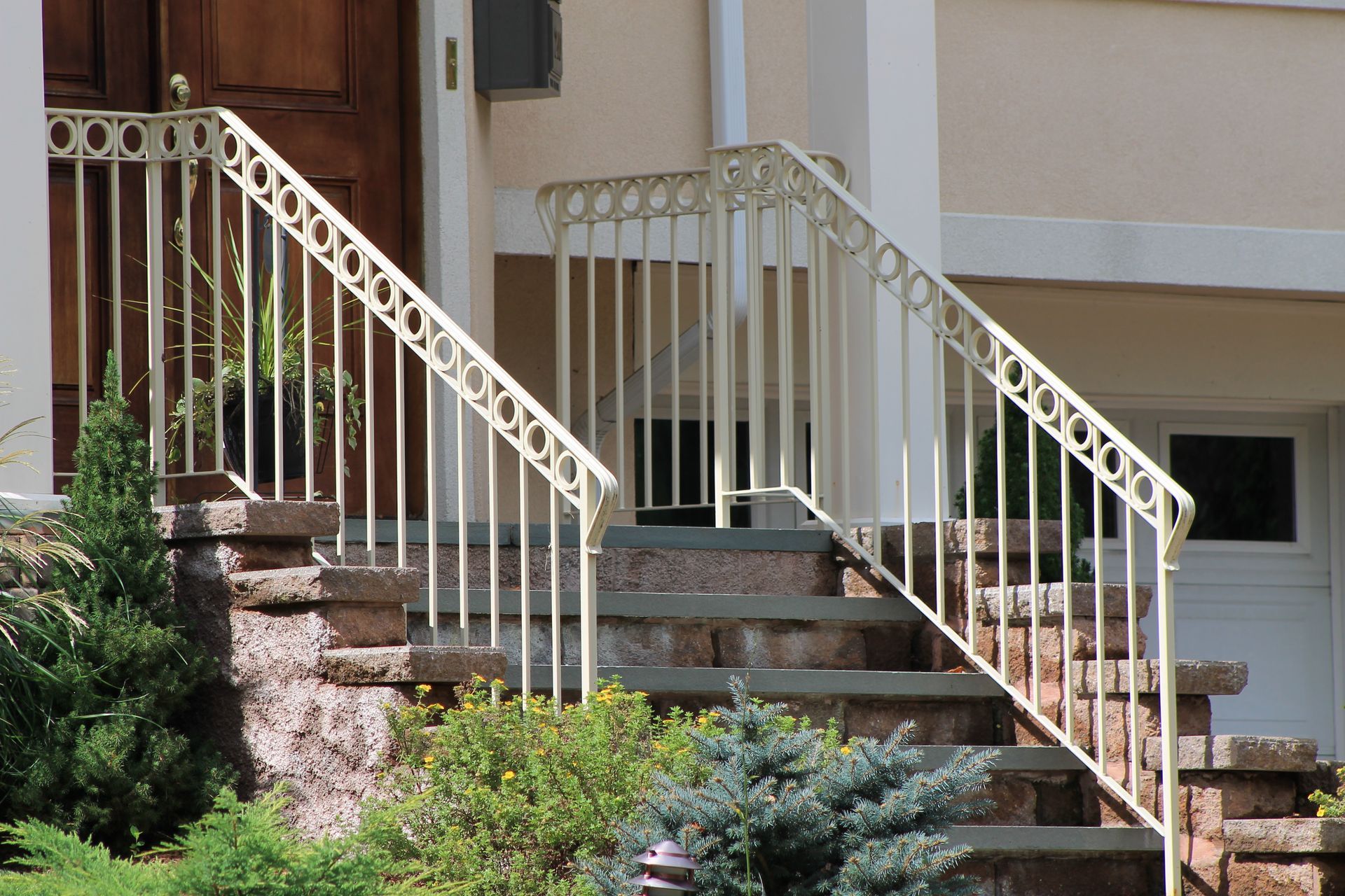 Front steps with white railings leading to a house entrance, bordered by shrubs and stone steps