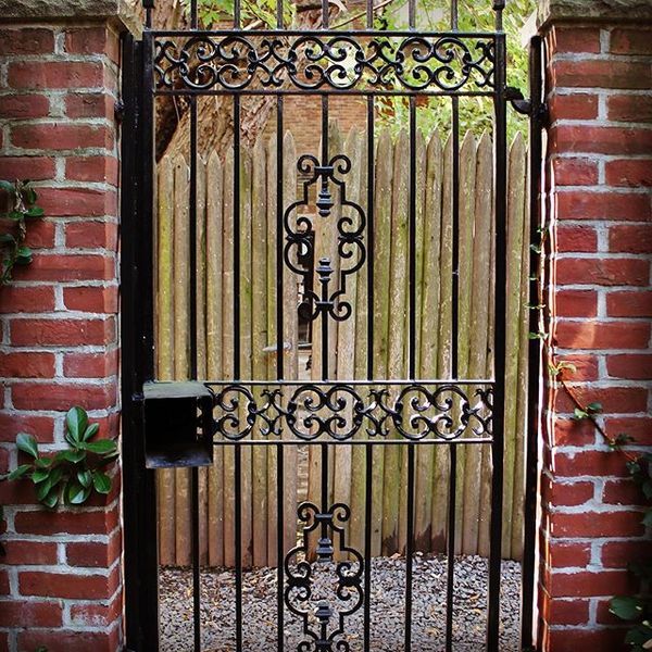 A black wrought-iron gate with decorative scrolls set between two brick pillars, in front of a wooden fence.