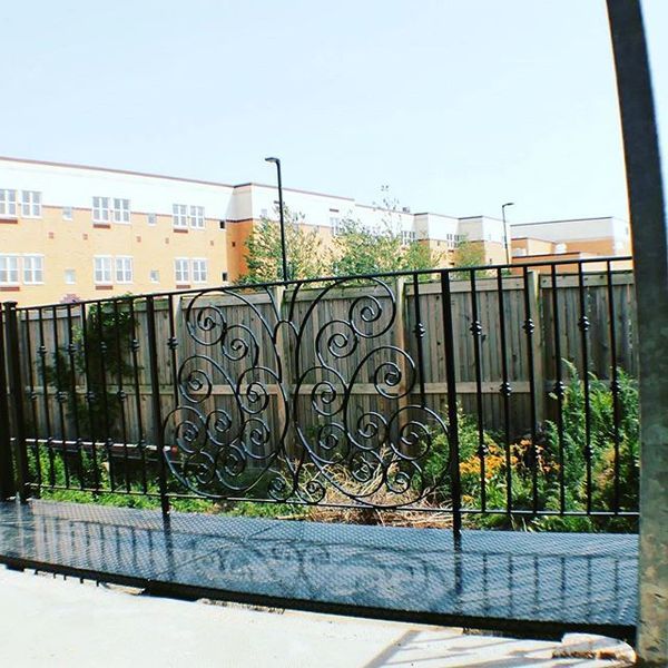 A black metal railing with decorative scrollwork stands before a wooden fence, with an apartment building in the distance.