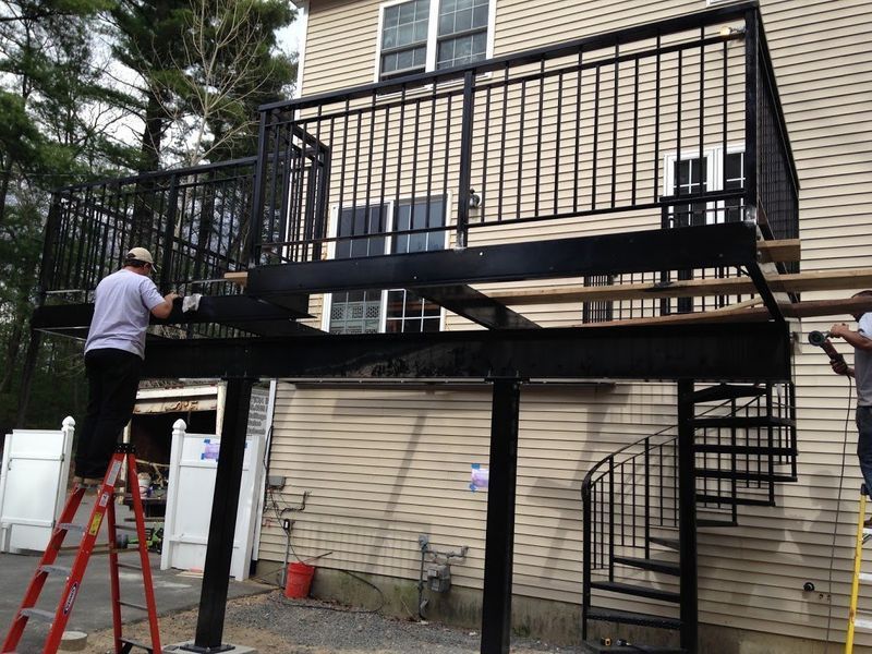 Two workers install a black metal deck and spiral staircase on the side of a two-story home.