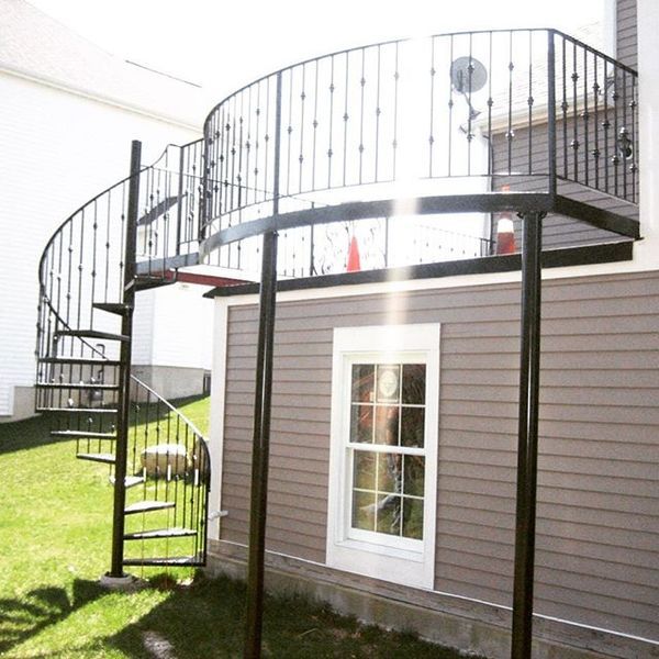A black metal spiral staircase leads up to a semi-circular raised deck attached to the side of a grey-sided house.