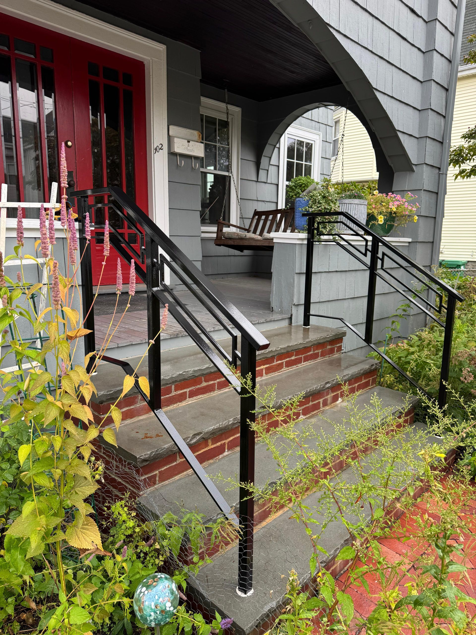 Front steps with black railings leading to a red door and arched porch at a gray house, with a small garden