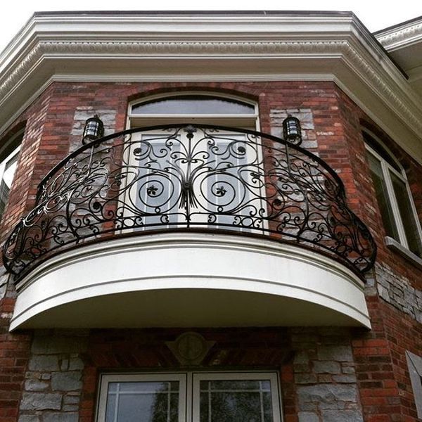 A curved balcony with ornate black wrought iron railings on a brick and stone house exterior with two wall sconces.