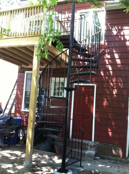 A black metal spiral staircase stands outdoors next to a red-sided building leading up to a wooden deck.