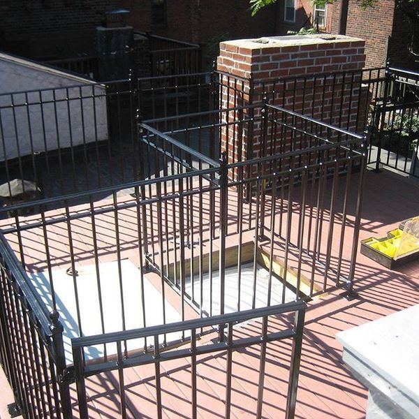 A rooftop deck features a black metal safety railing surrounding a square hatch opening next to a brick chimney.