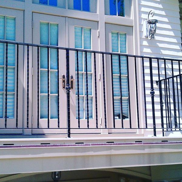 A black metal railing on a balcony in front of French doors with glass panes and light-colored siding.
