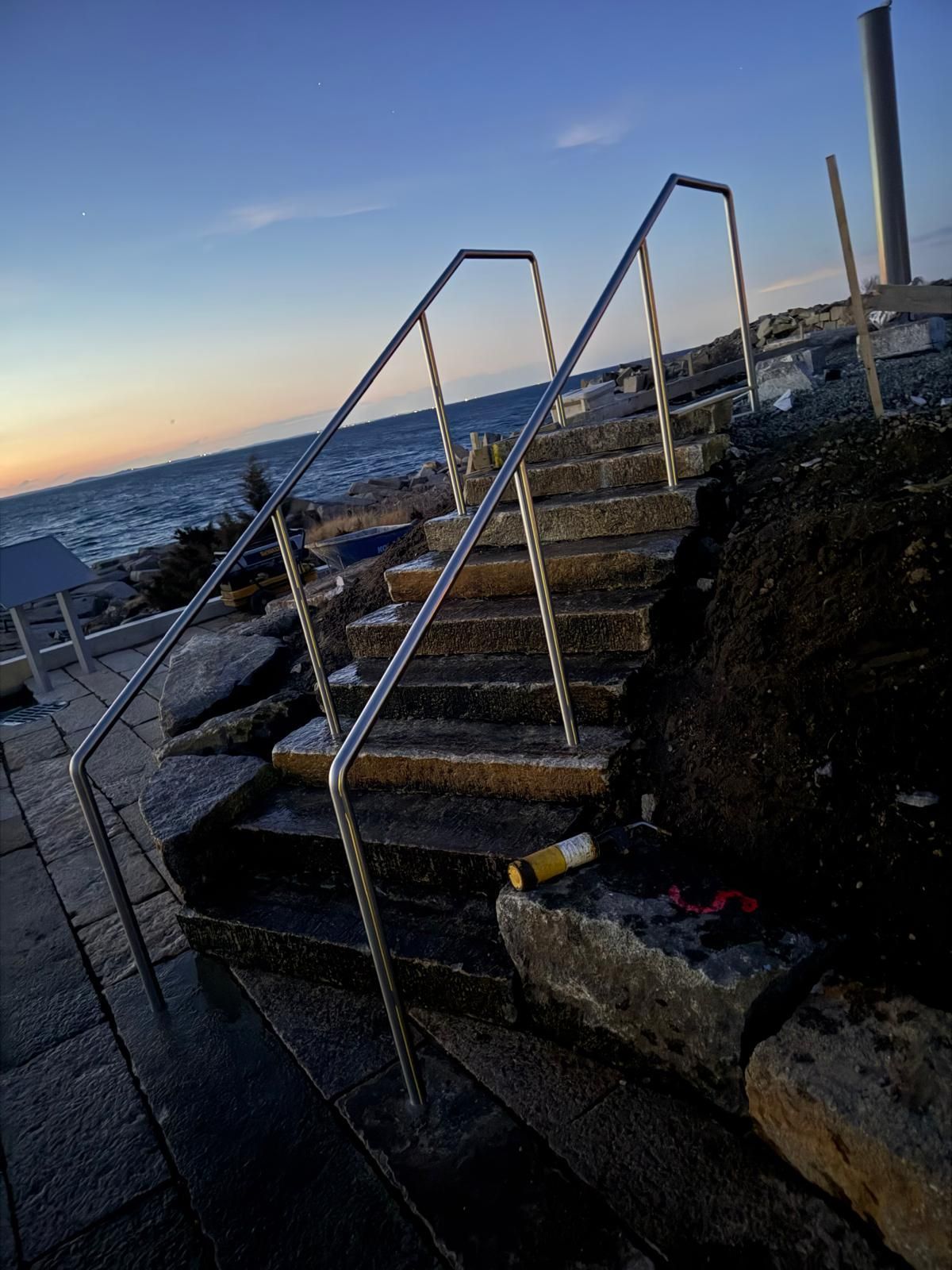 Metal stairs descending to a rocky shoreline at sunset, with ocean waves and a chain-link fence