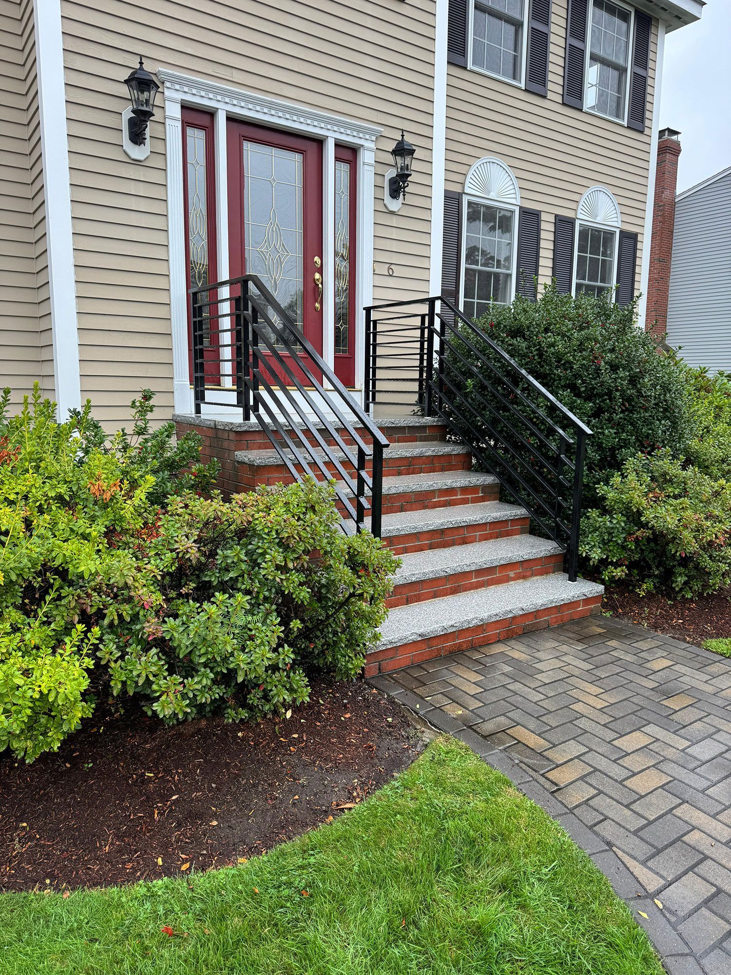 Front entrance of a townhouse with brick steps, black railings, red double doors, and landscaped shrubs