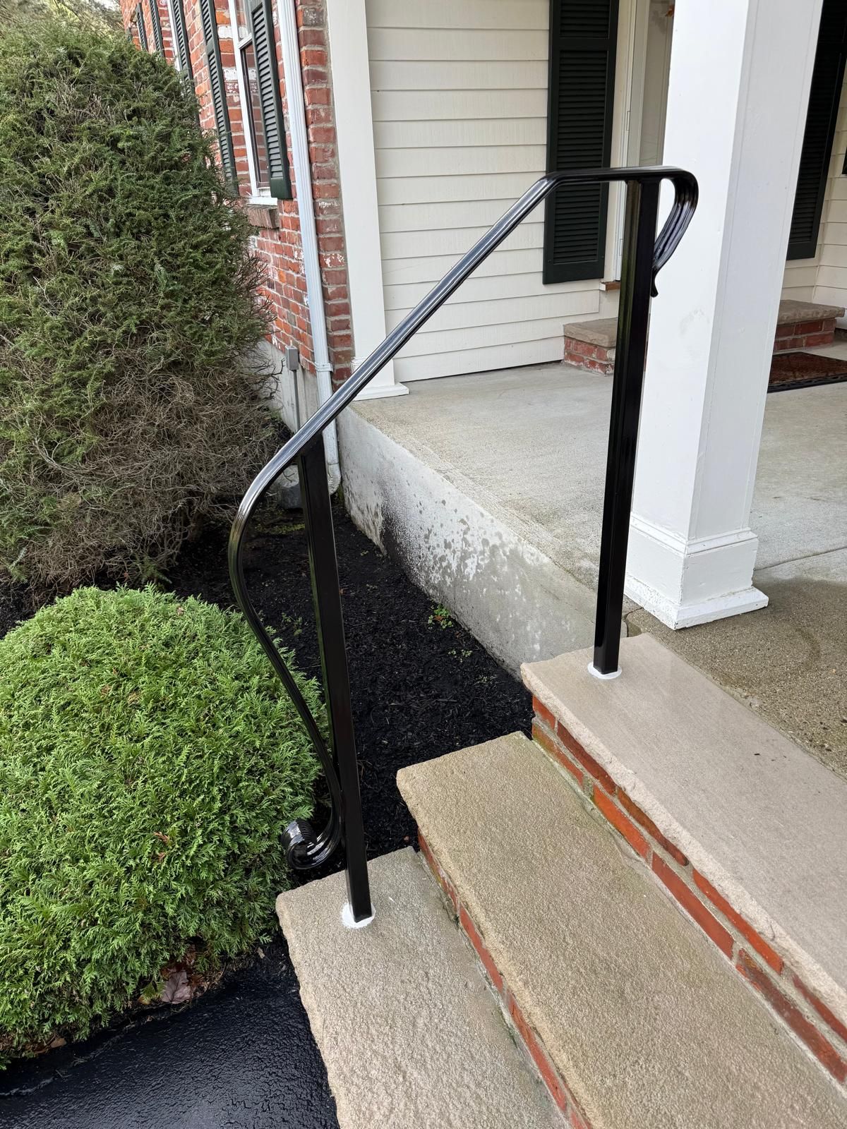 Black handrail beside concrete front steps and porch with shrubs and brick house exterior