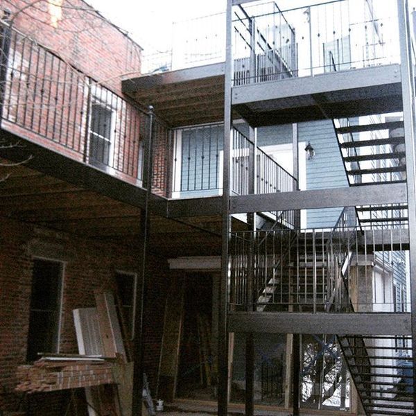 Multi-story steel exterior stairwell and wooden balcony attached to the side of a brick building under construction.