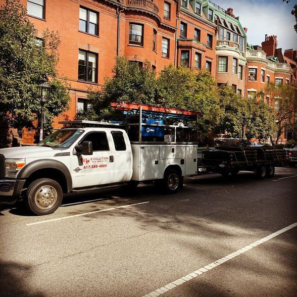 A white utility truck with a ladder rack and attached trailer parked on a street in front of red brick apartment buildings.
