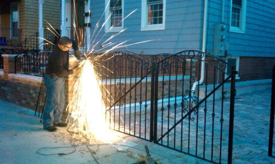 A person uses an angle grinder to cut a black metal fence, creating a bright spray of sparks on a sidewalk.