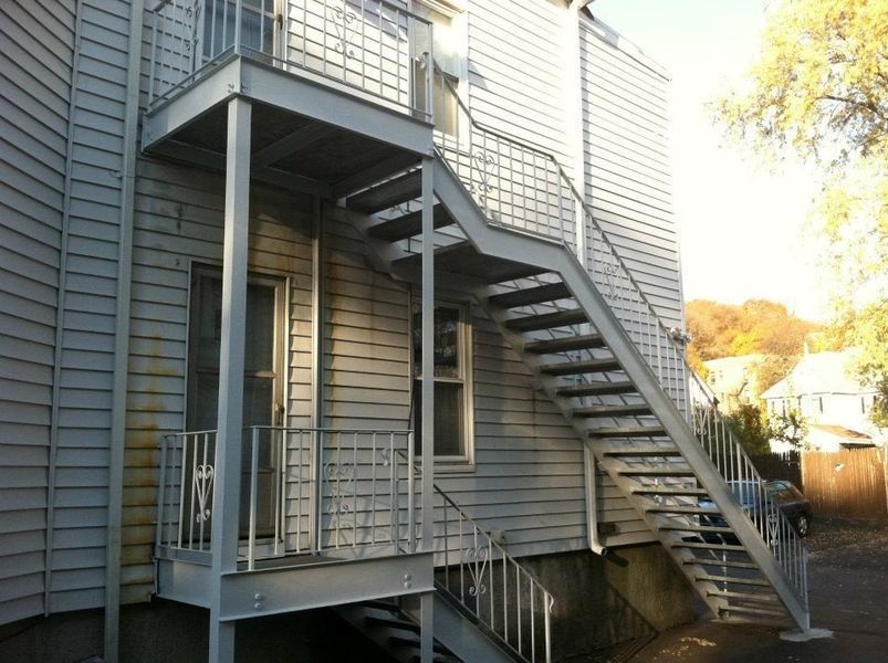 A light-gray metal exterior staircase attached to the side of a multi-story house with light-colored siding.