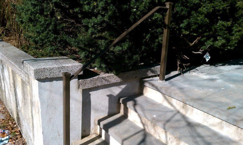 Concrete outdoor stairs with a single brown metal handrail leading up to a landing next to a stone retaining wall.