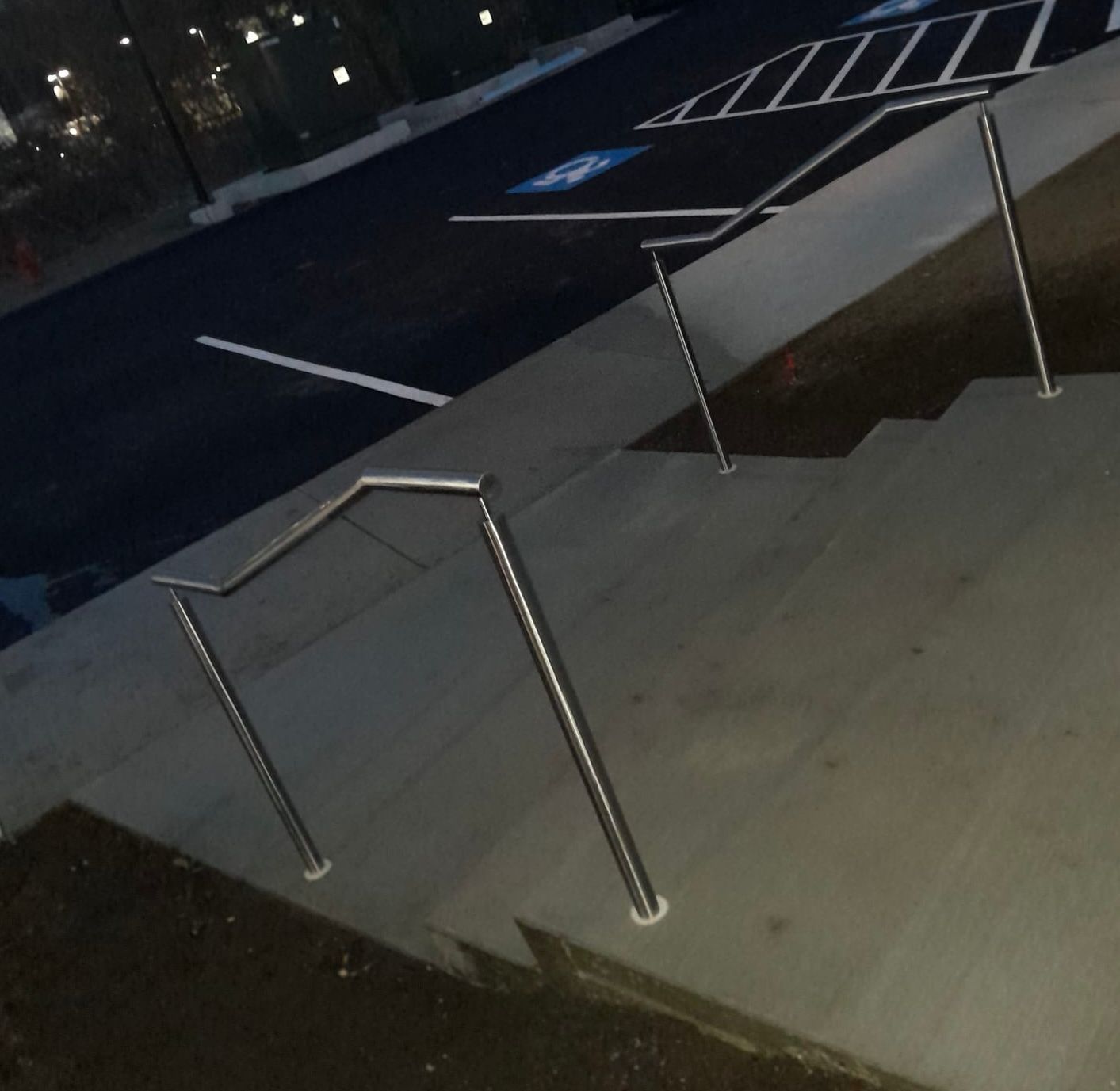 Nighttime sidewalk with metal railings beside a sloped curb ramp and parking lot markings