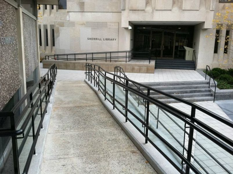 A concrete wheelchair ramp with black metal railings leading to the entrance of the Enigma Library building.