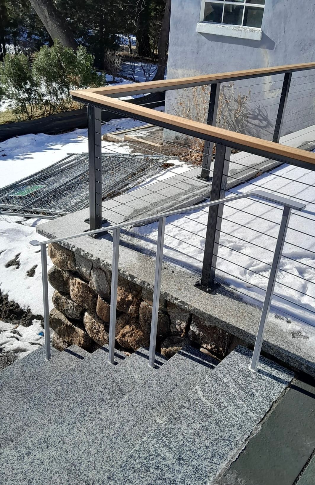 Exterior ramp with metal railings and a wooden handrail beside a gray building, above snow-covered ground
