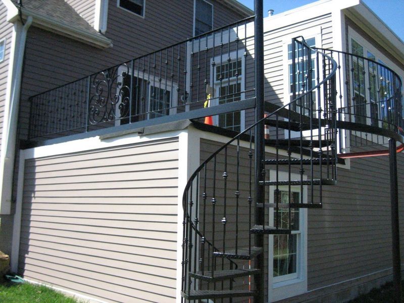 A multi-story house with tan horizontal siding features an outdoor deck with black metal railing and a spiral staircase.