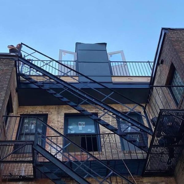 A rear exterior view of a brick building featuring black metal fire escapes and a rooftop deck with open glass doors.