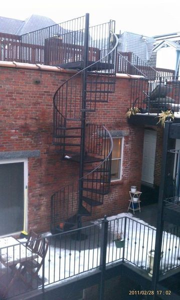 Black metal spiral staircase mounted on a brick building exterior, leading to a rooftop deck with metal railings.