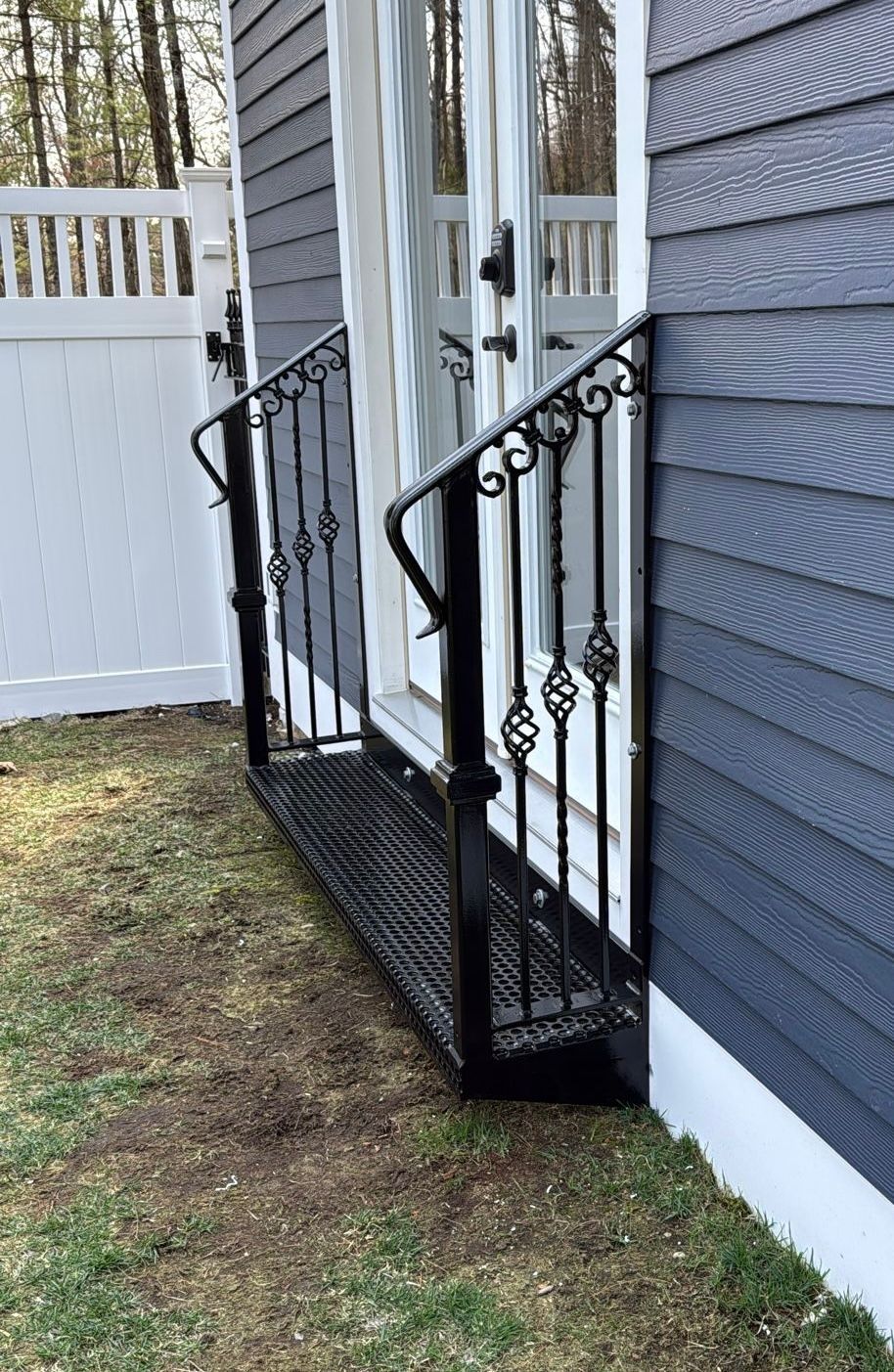 Black metal porch steps beside a gray house and white fence, with a black landing platform on grass