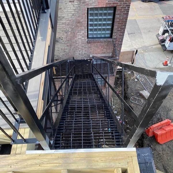 A high-angle view of a black metal outdoor staircase leading down alongside a brick building with a glass block window.