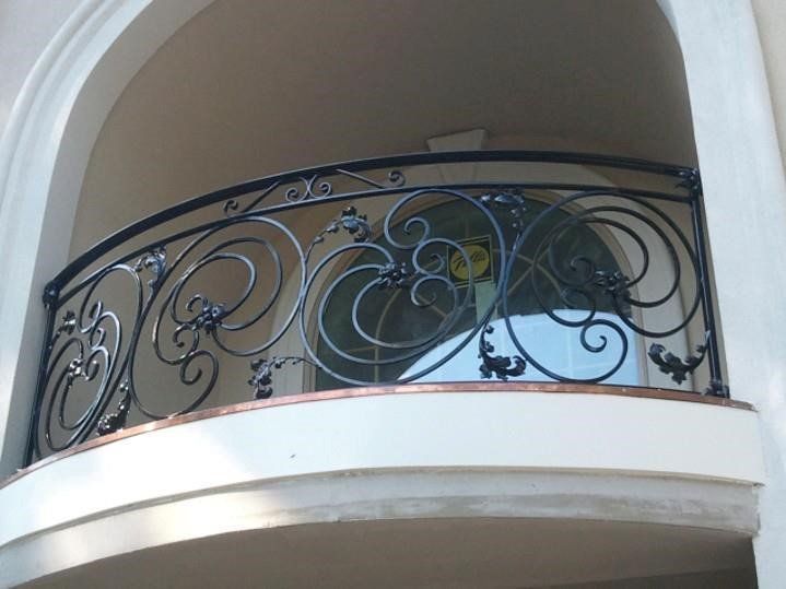 A curved black wrought-iron railing with ornate scrollwork, installed on a balcony set within a stone-arched alcove.