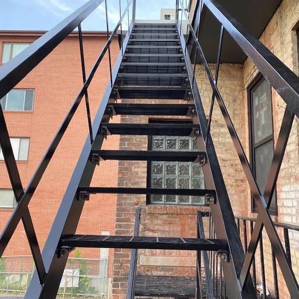 A steep, black metal outdoor fire escape staircase leading upward against a brick building.