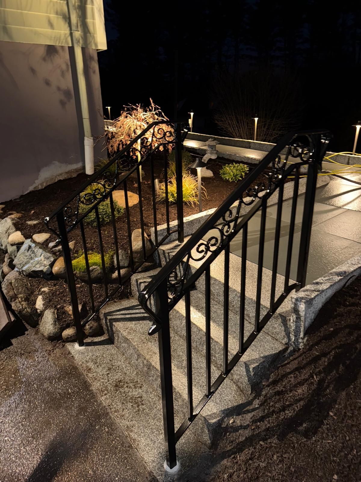 Nighttime outdoor stone steps with black iron railings beside a landscaped rock garden