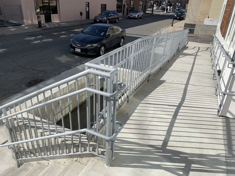 A view from a concrete staircase looking toward an accessible ramp with white metal railings next to a city street.