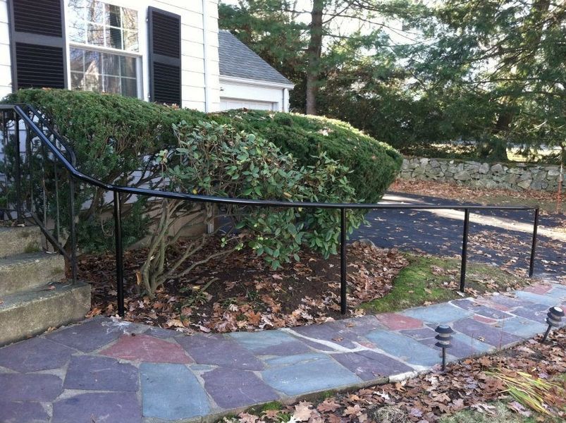 A black metal handrail runs along a stone walkway and garden edge next to the steps of a white house.