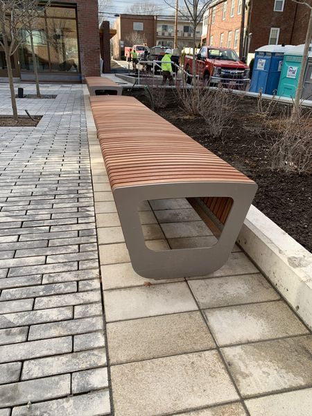 Modern wooden-slatted bench with gray metal supports along a paved path next to a planting bed and buildings.