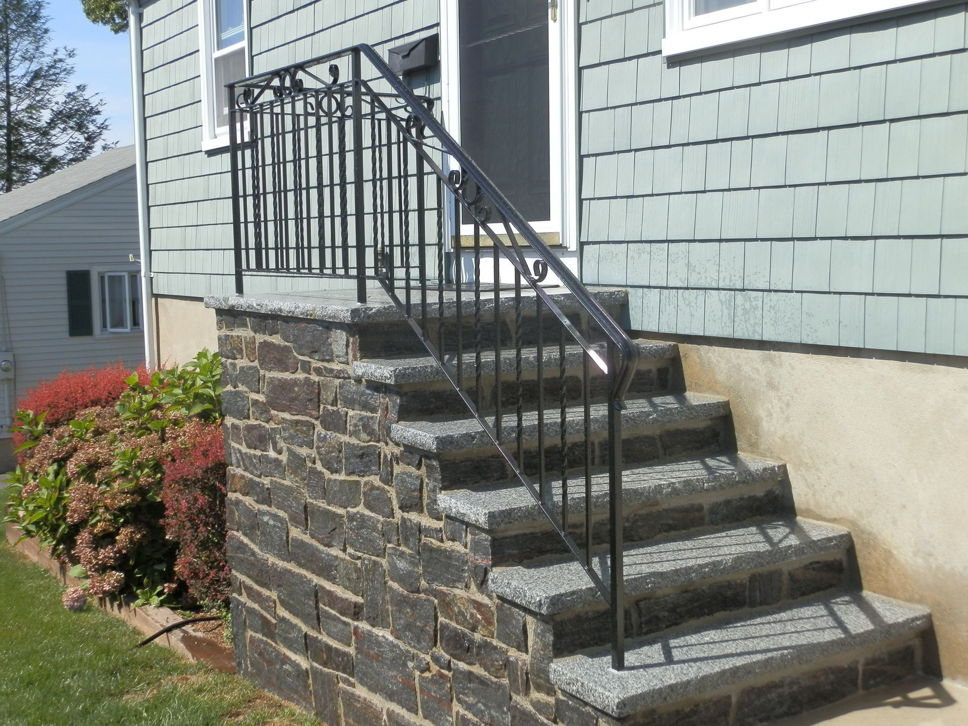Front steps with stone retaining wall and black metal railing leading to a house entrance