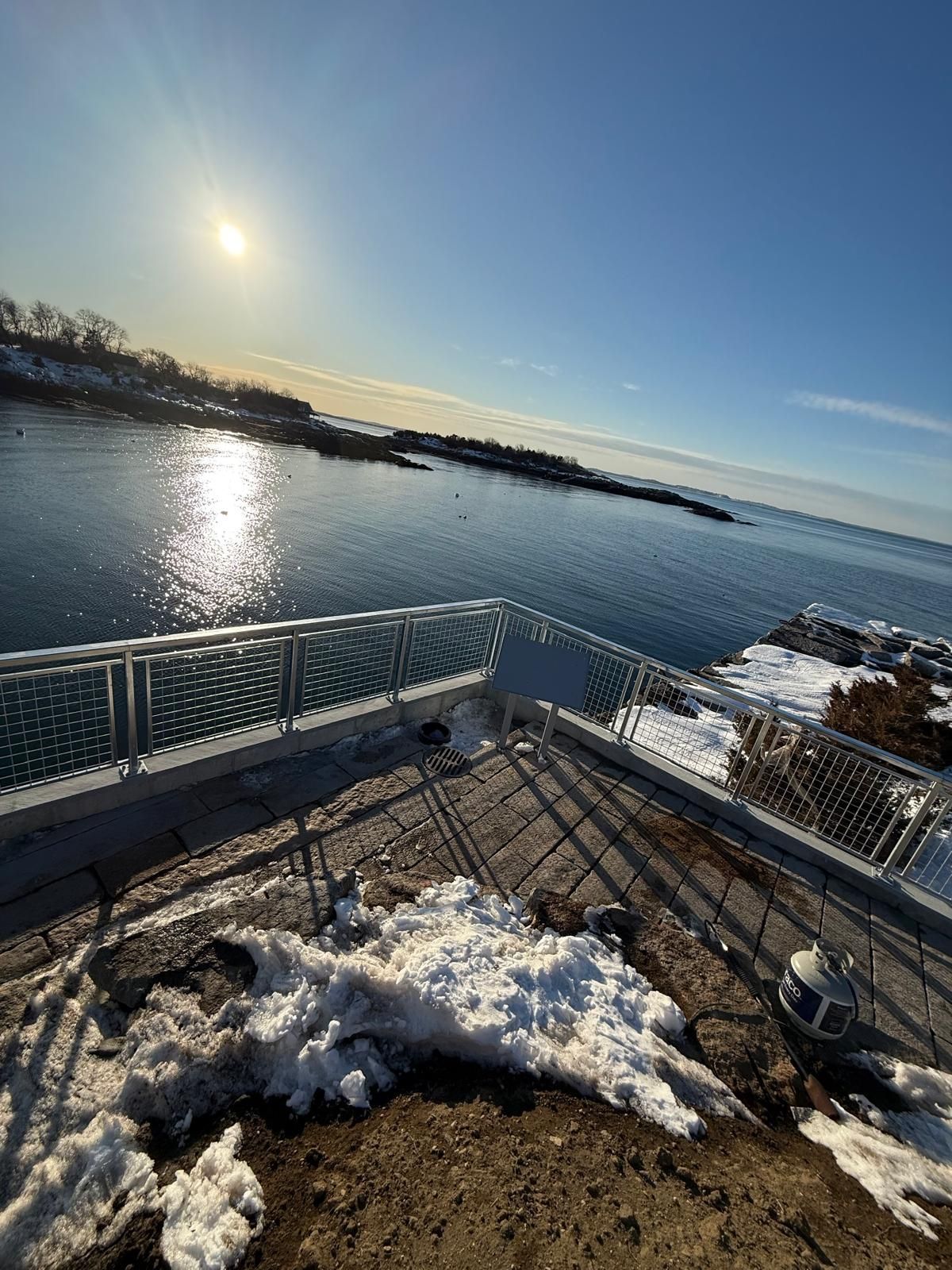 Snowy waterfront deck overlooking calm blue water at sunrise, with icy railings and a bright sky