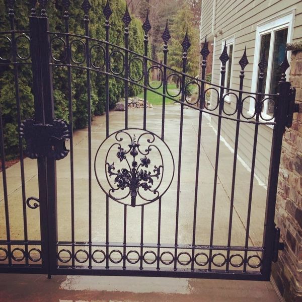 A black iron gate with a decorative circular floral centerpiece, positioned in front of a house with light siding.