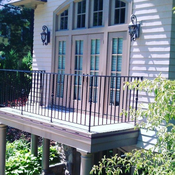 A beige house with a raised wooden balcony featuring black metal railings, leading to arched glass double doors.