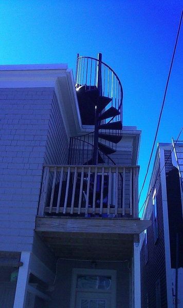 A black spiral staircase ascends from a wooden balcony to the roof of a light-colored building under a clear blue sky.