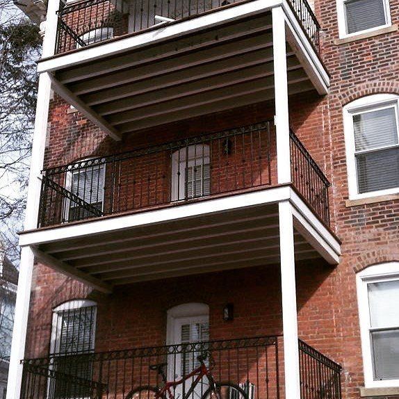 A red brick building with three stacked balconies, white support pillars, and a bicycle on the lowest level.