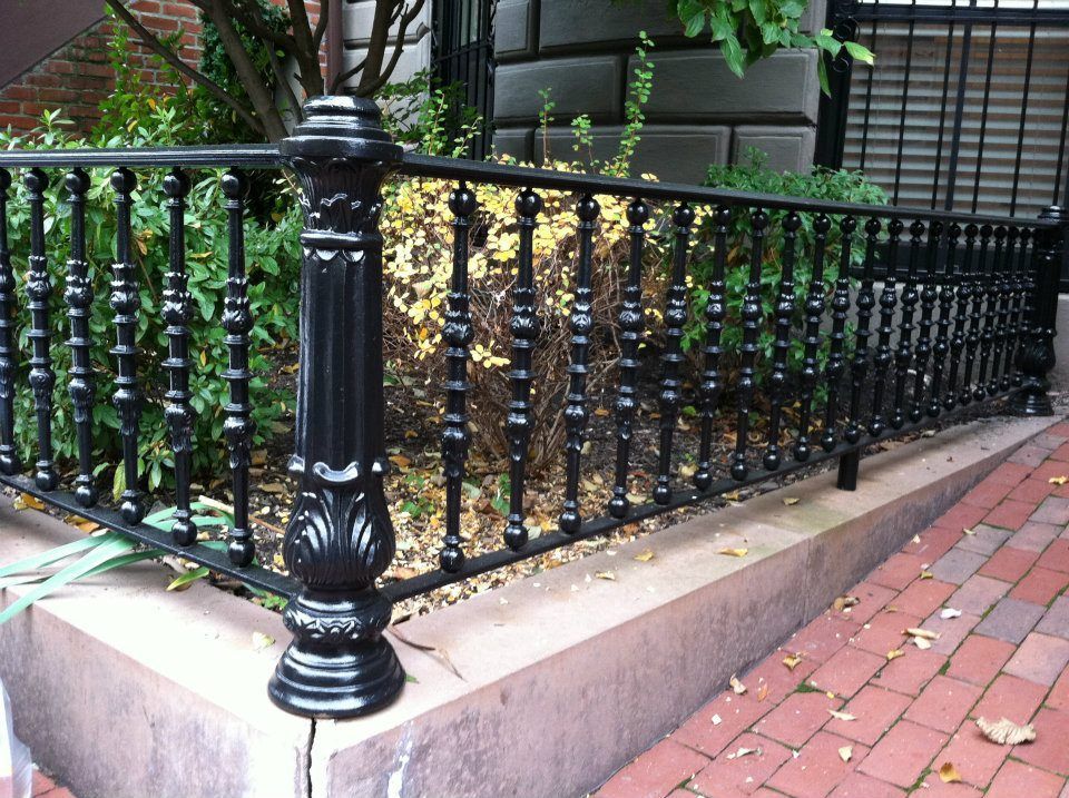 Black wrought-iron fence around a small garden beside a red brick sidewalk