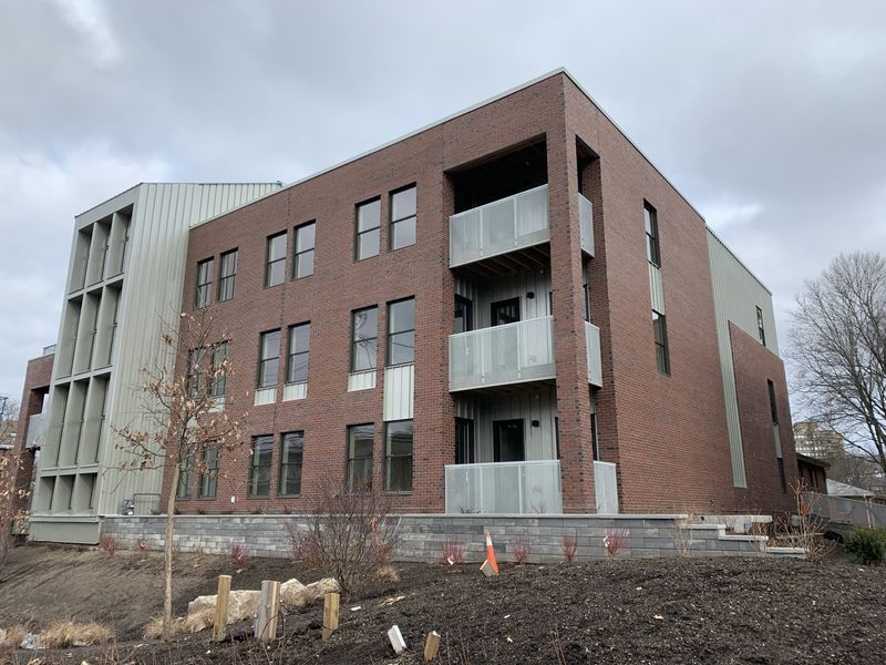A modern three-story brick building with metal siding and balconies, set against a cloudy sky.