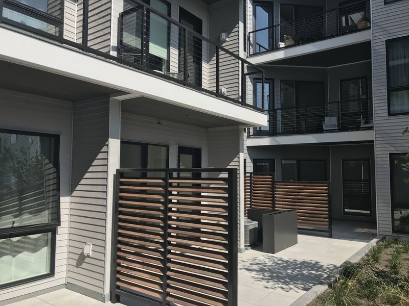 Modern apartment complex exterior with light gray siding, private patios, horizontal wooden privacy screens, and balconies.