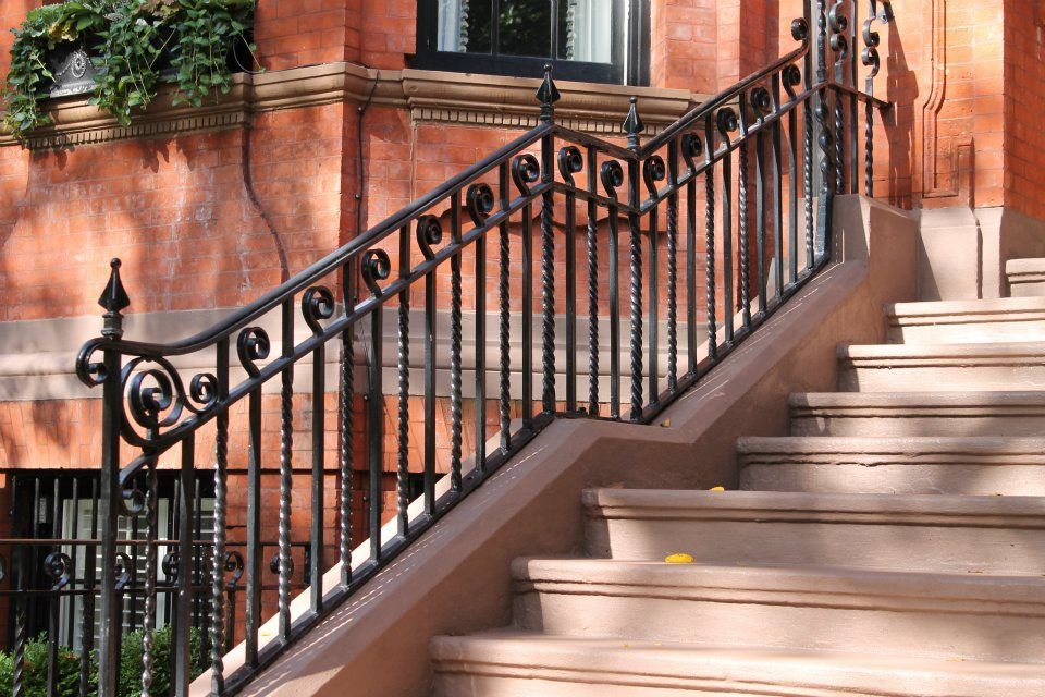 Staircase with ornate black iron railing beside a red brick building and potted greenery