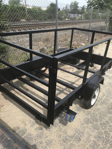 A black, rectangular utility trailer with metal rail sides, parked on a paved surface near a chain-link fence.