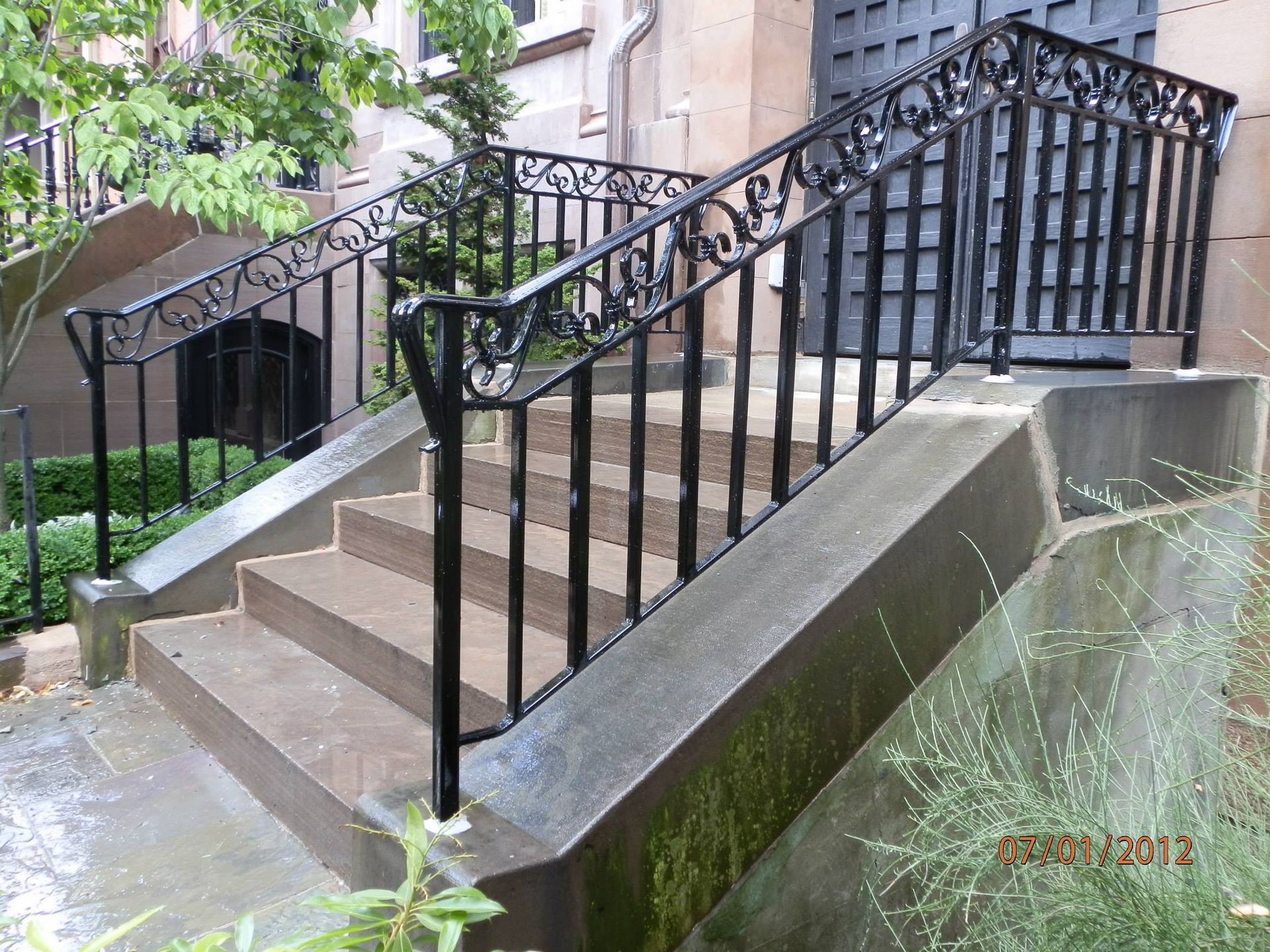 Outdoor stairway with black metal railings beside a wet concrete wall and building steps